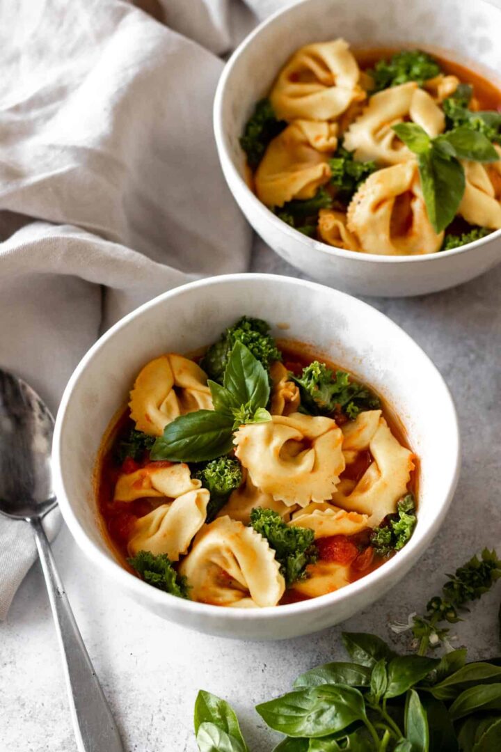 two bowls served with tortellini soup with kale, tomatoes and garnished with fresh basil