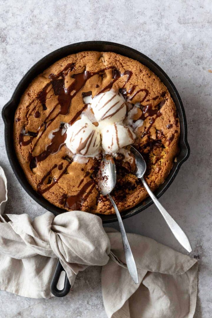 a black skillet with chocolate chip cookie and three ice cream balls on top and chocolate topping