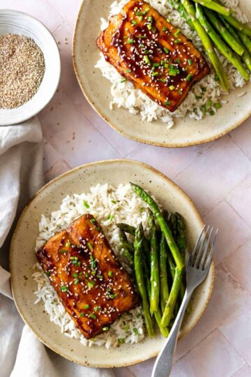 two plates served with white rice, baked salmon, asparagus, chopped chives, sesame seeds and a small bowl with sesame seeds on the side