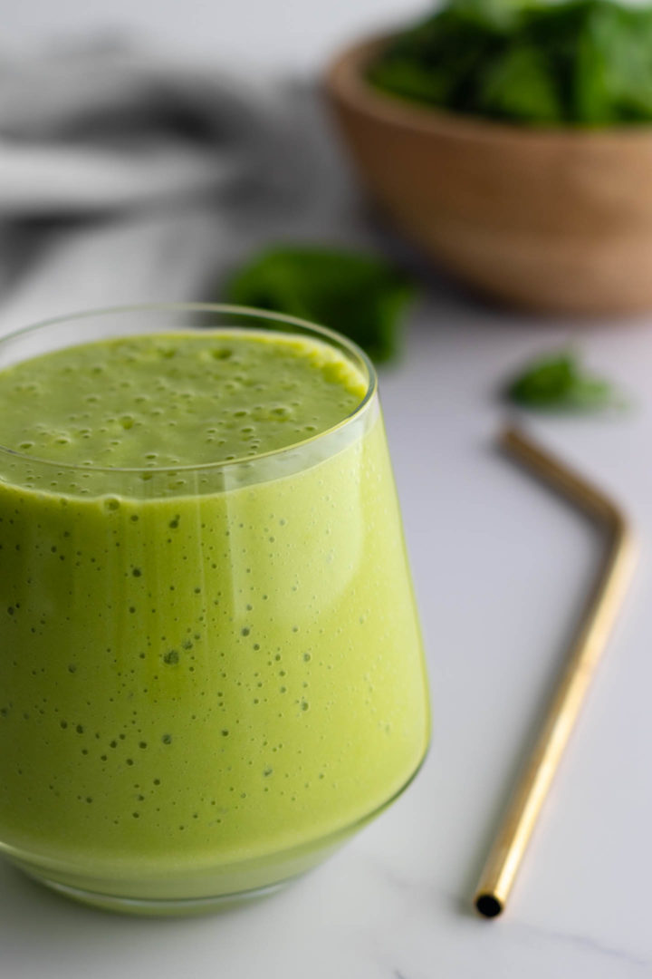 a glass with green smoothie, a golden straw and a bowl with spinach out of focus