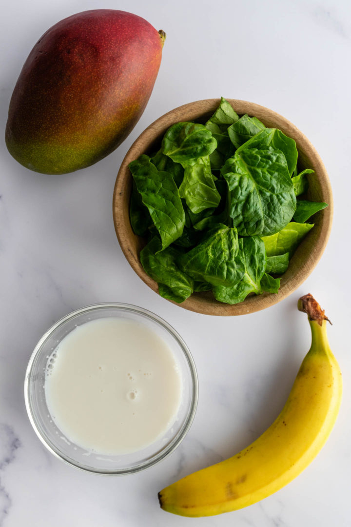 one mango, a bowl with spinach leaves, a banana and a bowl with almond milk