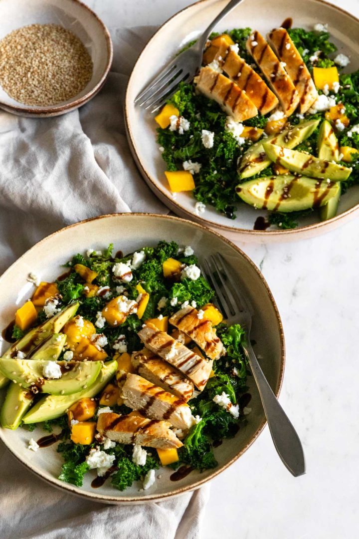 two plates served with kale, avocado, chicken, mango, feta cheese, sesame seeds and balsamic glaze. One small bowl with sesame seeds