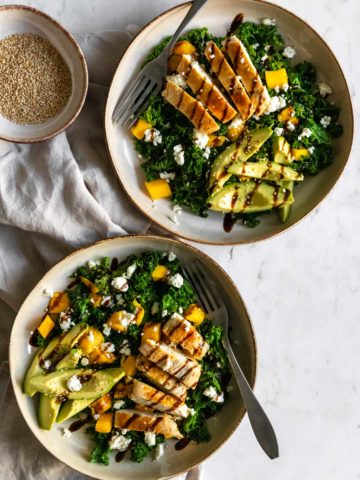 two plates with kale, chicken, avocado, feta cheese, mango, sesame seeds, and a bowl with sesame seeds on the side