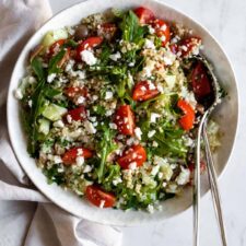a bowl with quinoa salad on top of a marble table, two serving spoons and a kitchen towel