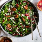 a large bowl with kale, arugula, pecan nuts, feta cheese, pomegranate seeds and two serving spoons. two small bowls on the side, one with pecans the other one with crumbled feta and pomegranate