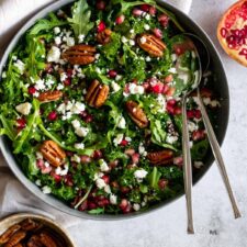 a large bowl with kale, arugula, pecan nuts, feta cheese, pomegranate seeds and two serving spoons. two small bowls on the side, one with pecans the other one with crumbled feta and pomegranate