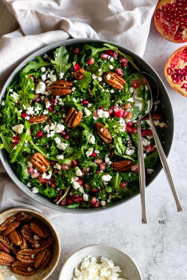 a large bowl with kale, arugula, pecan nuts, feta cheese, pomegranate seeds and two serving spoons. two small bowls on the side, one with pecans the other one with crumbled feta and pomegranate