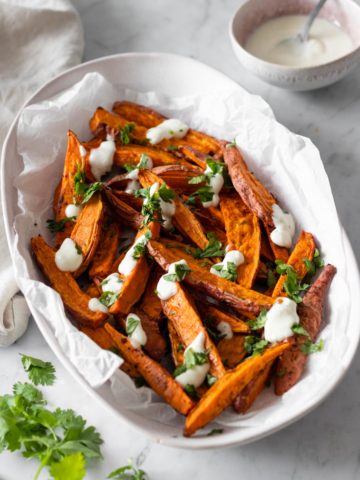 sweet potato wedges on a plate with chopped coriander and garlic dressing. A small bow with more garlic dressing on the side