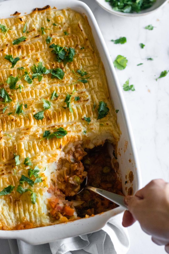 a baking dish with lentil Shepherd's pie, chopped coriander and a hand holding a spoon