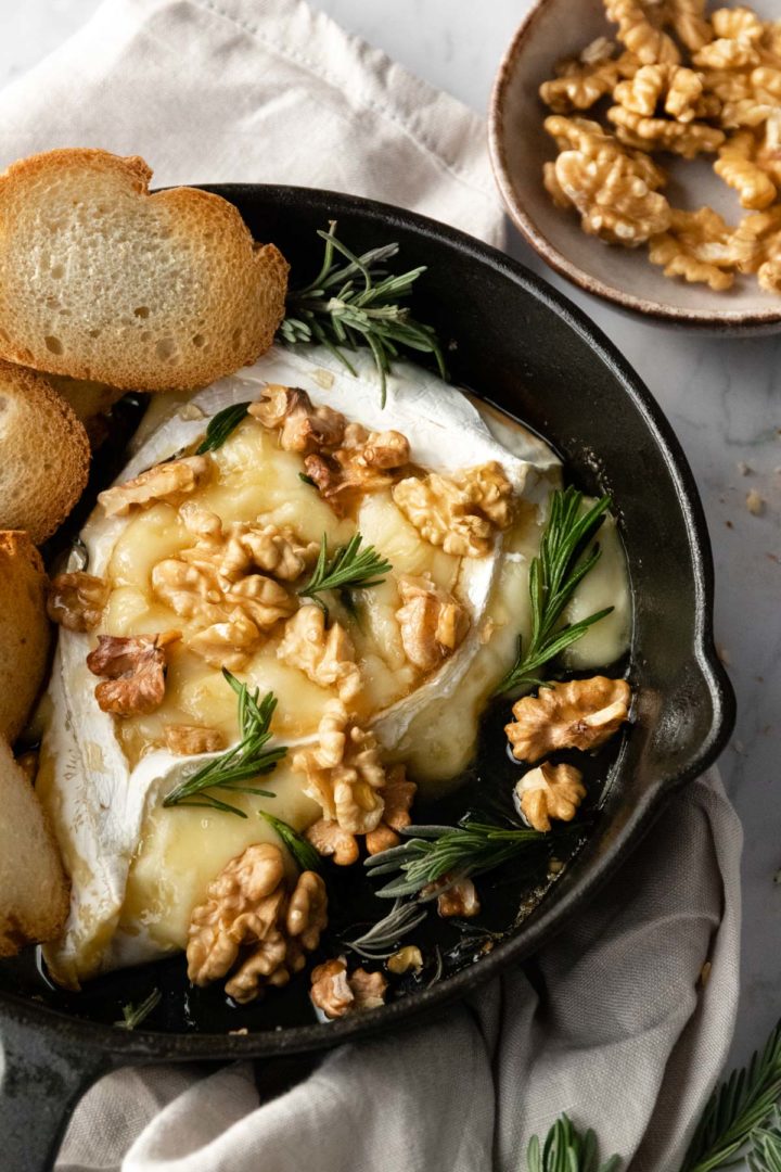 close-up of a skillet with baked brie and toasted bread with rosemary and walnuts on top