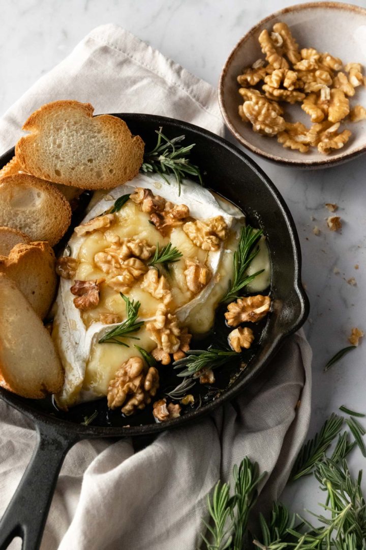 A skillet with baked brie with walnuts, rosemary, toasted bread and a small bowl with more walnuts on the side