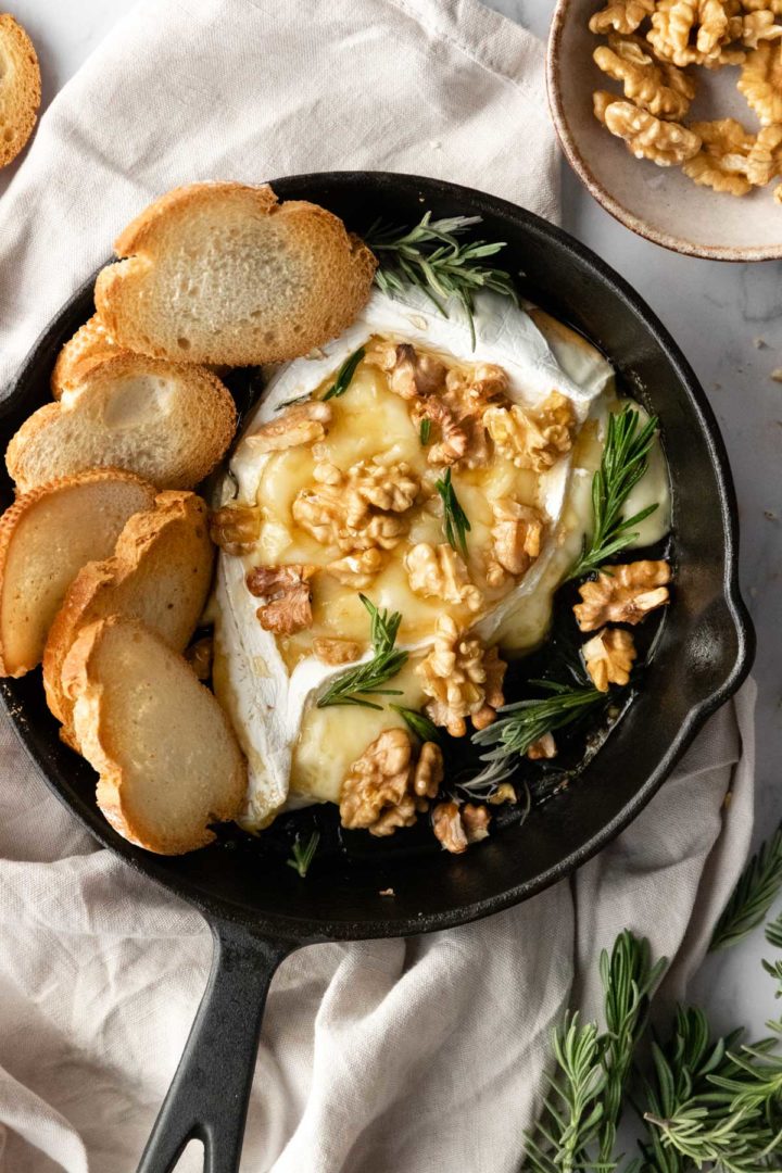 baked brie, toasted bread, walnuts and rosemary inside a black skillet, more rosemary and walnuts on the side
