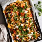 a baking tray with chicken sheet pan nachos topped with sour cream and coriander, a small bowl with coriander