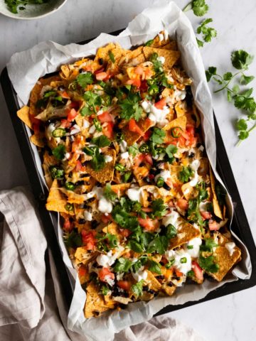 a baking tray with chicken sheet pan nachos topped with sour cream and coriander, a small bowl with coriander