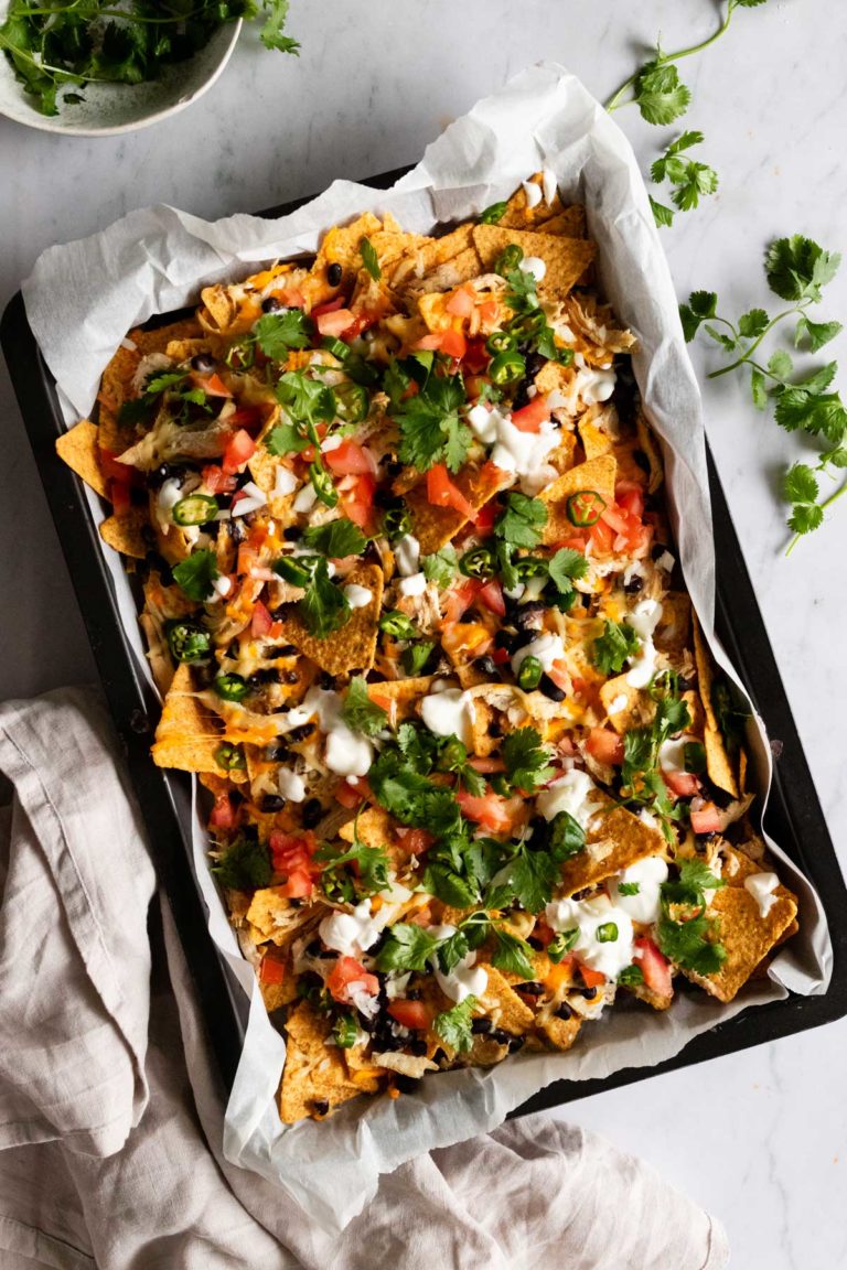 a baking tray with chicken sheet pan nachos topped with sour cream and coriander, a small bowl with coriander