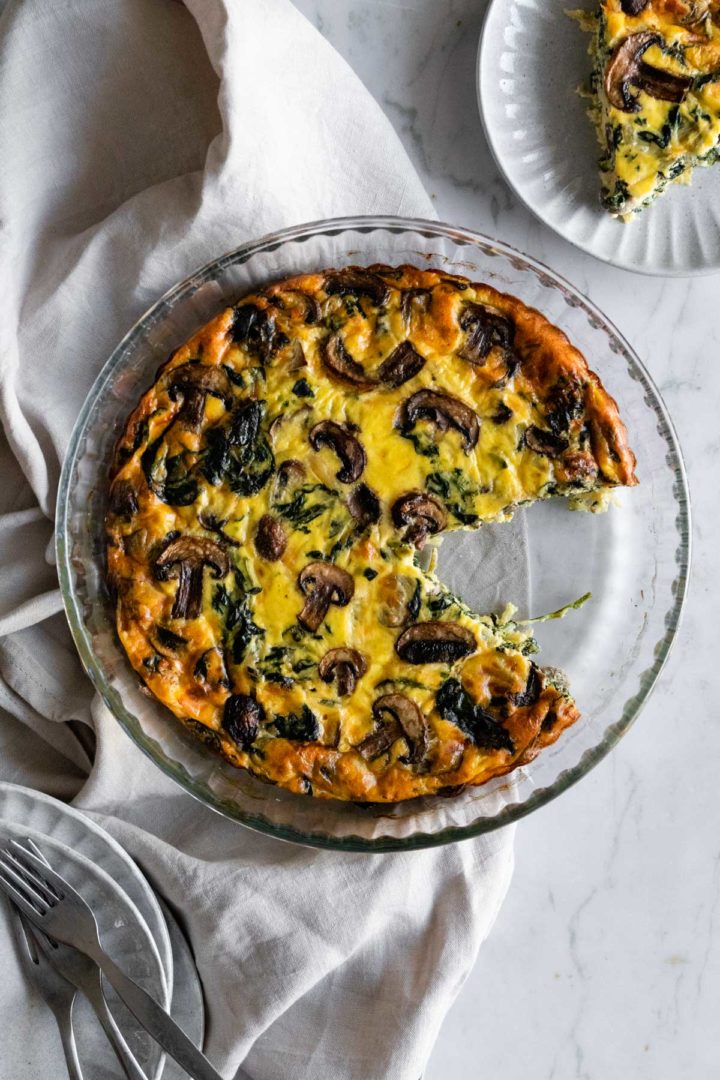 one quiche with mushrooms and spinach on top of a kitchen towel, one plate with one quiche slice on the top right corner