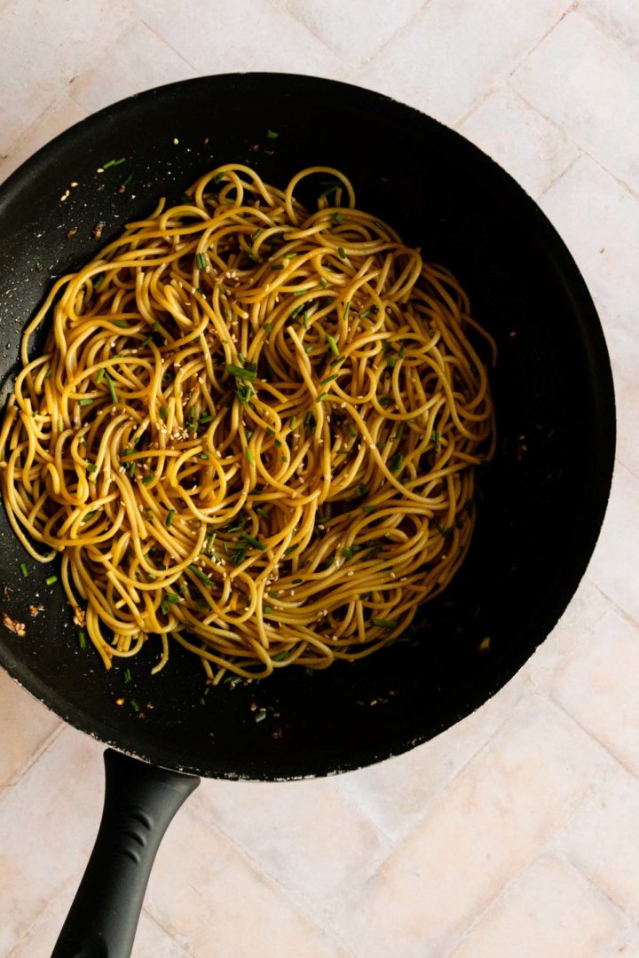 sesame noodles inside a wok with sesame seeds and chopped chives on top