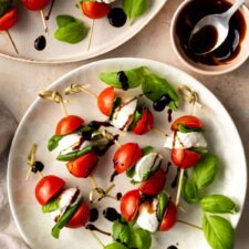 two plates served with caprese skewers garnished with blasamic glazed and basil leaves. A small bowl with a spoon and more balsamic glaze on the side