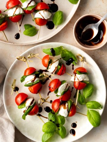 two plates served with caprese skewers garnished with blasamic glazed and basil leaves. A small bowl with a spoon and more balsamic glaze on the side