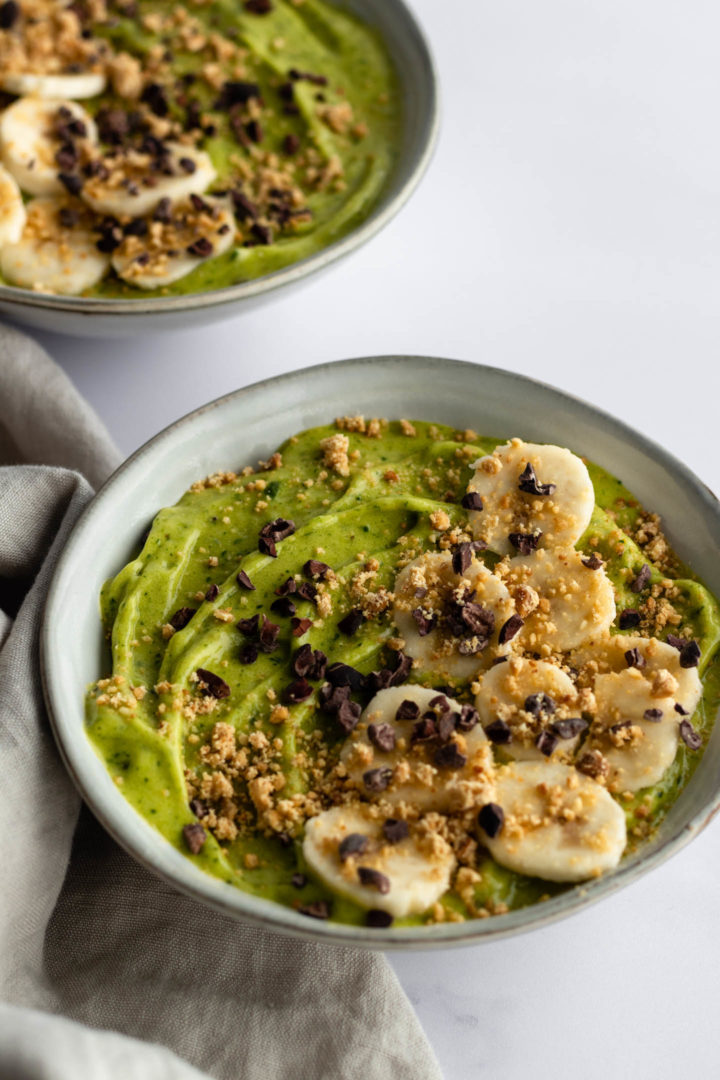 two bowls served with green smoothie, sliced banana, cacau nibs and peanut crumble