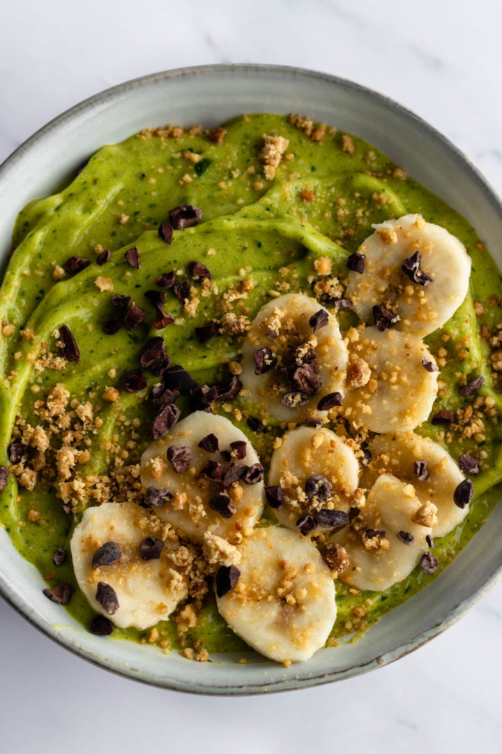 close up of a breakfast bowl with green smoothie, sliced banana, cacau nibs and peanut crumble