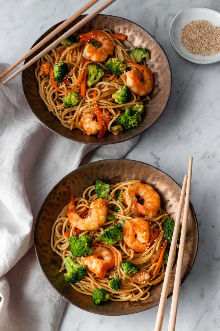 two bowls with noodles, shrimp, carrots, broccoli and sesame seeds. Chop sticks on each bowl and a small bowl with sesame seeds