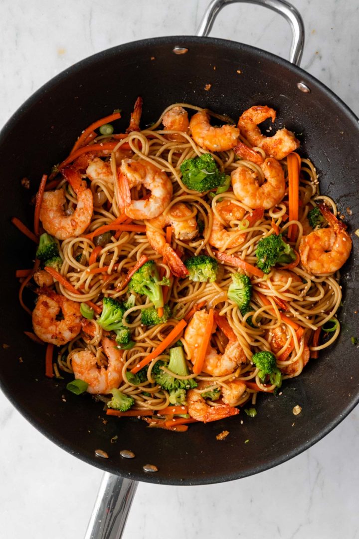 a large wok with noodles, shrimp, carrot, broccoli and sesame seeds on top