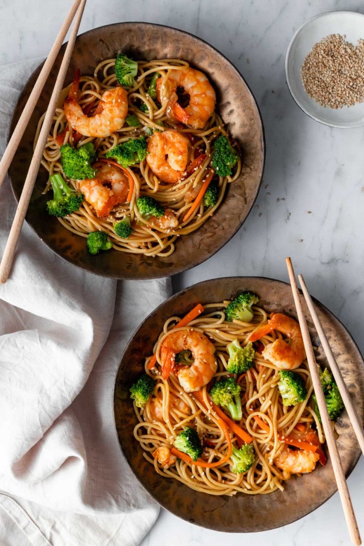 two bowls served with shrimp noodle stir fry, chop sticks and a small bowl on the side with sesame seeds