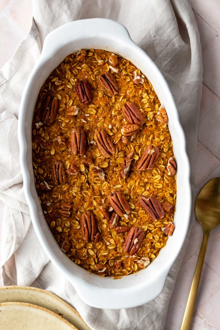 carrot cake baked oatmeal on a baking dish with pecans on top, a gold spoon on the side