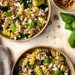 two plates served with pasta, zucchini, mushrooms, crumbled feta, spinach, walnuts and fresh basil. A small bowl with walnuts on the side
