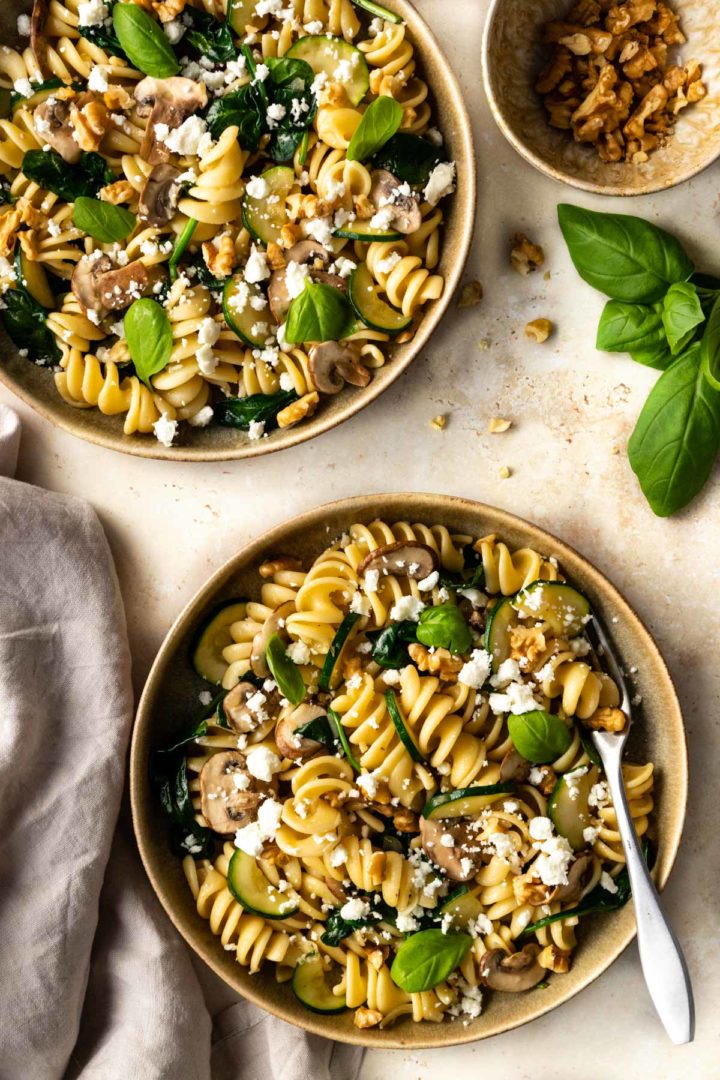 two plates served with pasta, zucchini, mushrooms, crumbled feta, spinach, walnuts and fresh basil. A small bowl with walnuts on the side