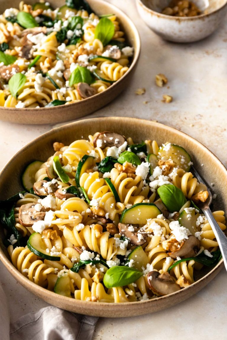 two plates served with pasta, zucchini, mushrooms, crumbled feta, spinach, walnuts and fresh basil. A small bowl with walnuts on the side
