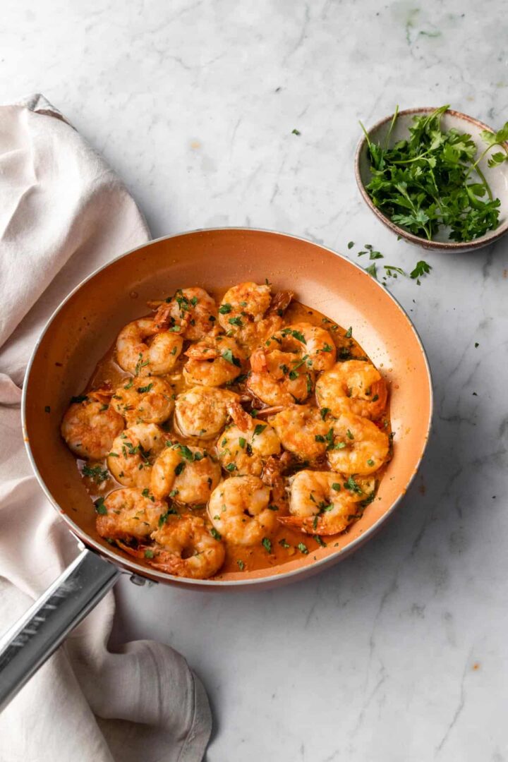 a pan with shrimp on top of a marble board with a kitchen towel and chopped parsley over the shrimp and on a small bowl on the side
