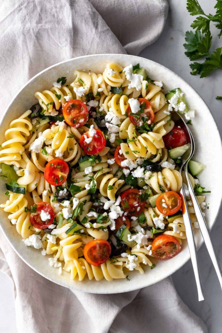 a large bowl with greek pasta salad with fusilli pasta, crumbled feta, cherry tomatoes, olives, cucumber and chopped parsley