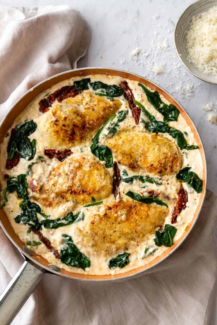 a pan with creamy Tuscan chicken with sun-dried tomatoes, baby spinach and a small bowl with parmesan cheese on the side