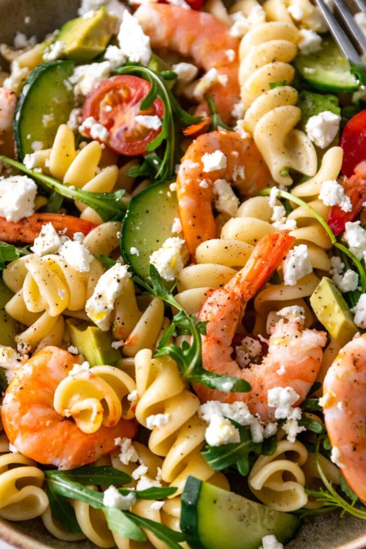 close up of a plate with shrimp, pasta, cucumber, arugula, feta cheese, avocado and cherry tomatoes