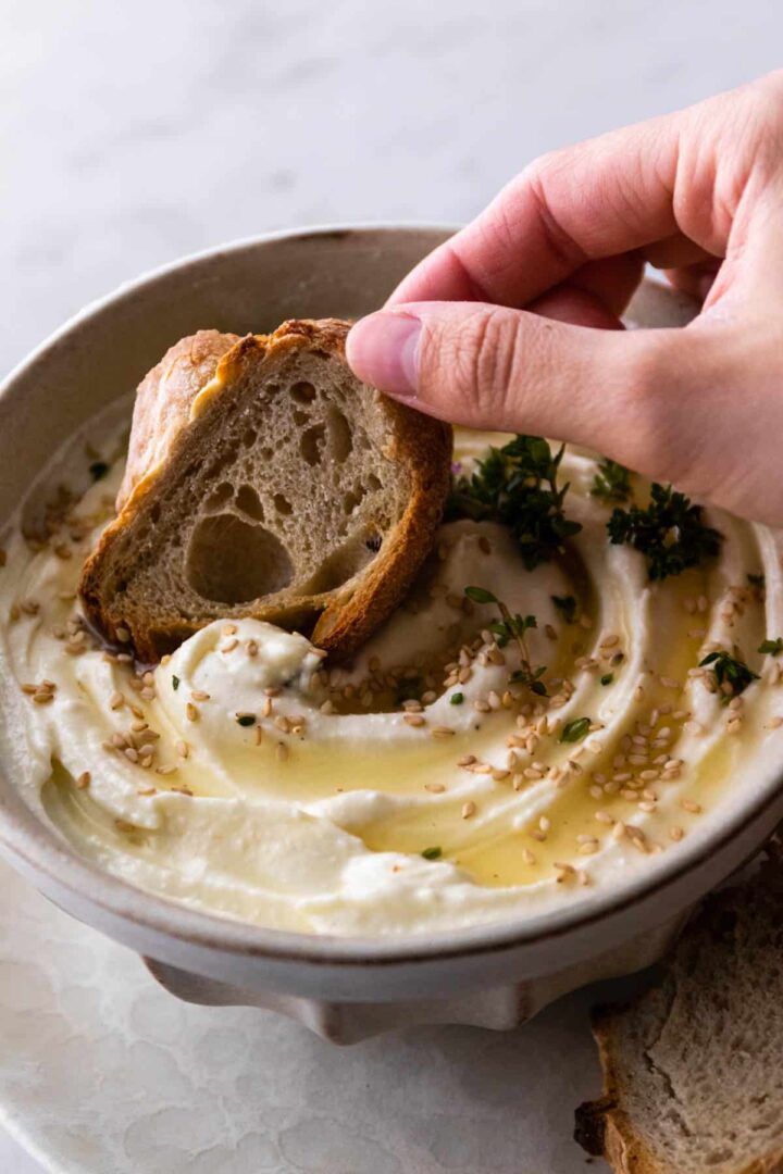 one hand holding a piece of toasted bread and dipping on the feta cheese