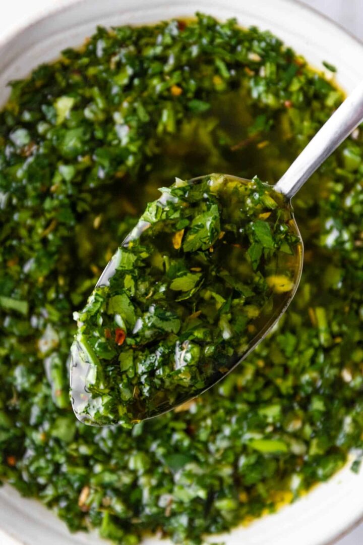 close up of a spoon with chimichurri sauce and a bowl with more sauce on the background