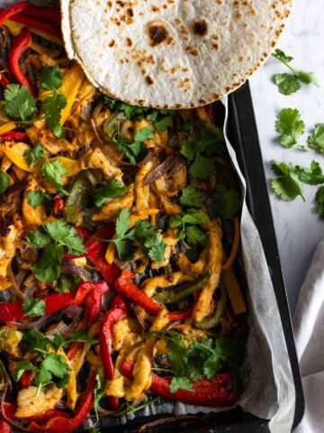 a baking tray with sheep pan chicken fajitas, aka chicken breast slices, bell pepper slices, sliced onion, coriander, and tortillas