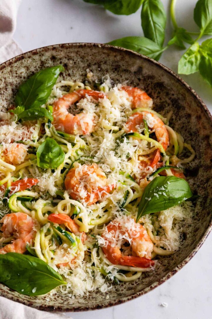 a bowl with zucchini noodles with shrimp garnished with grated parmesan cheese and basil leaves