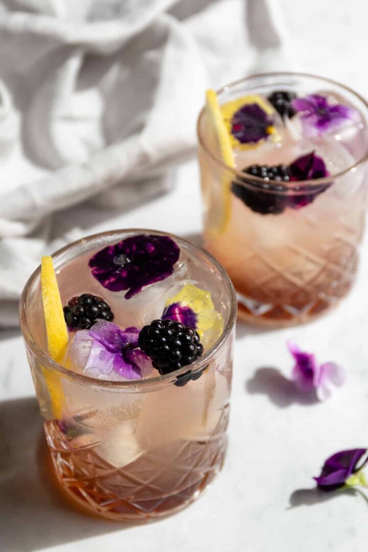 two glasses with bramble cocktail, lemon slice, blackberries, and flower ice cubes