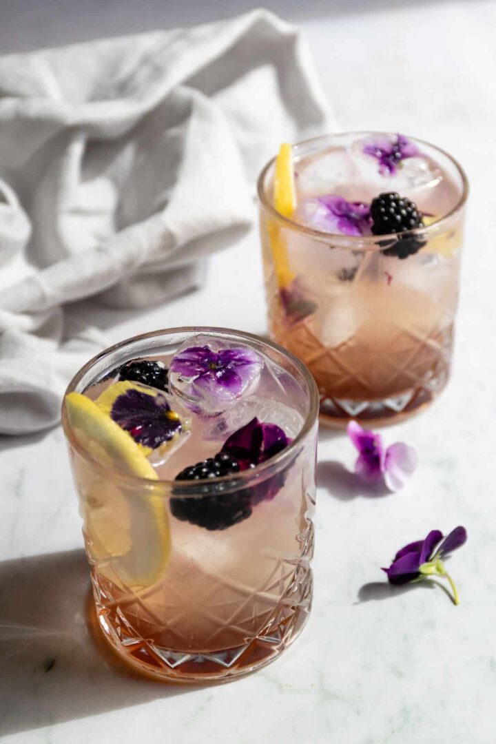 two glasses served with cassis bramble cocktail with lemon slice, flower ice cubs, and blackberries
