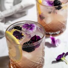 two glasses with bramble cocktail, wildflower ice cubes, lemon slice and blackberries