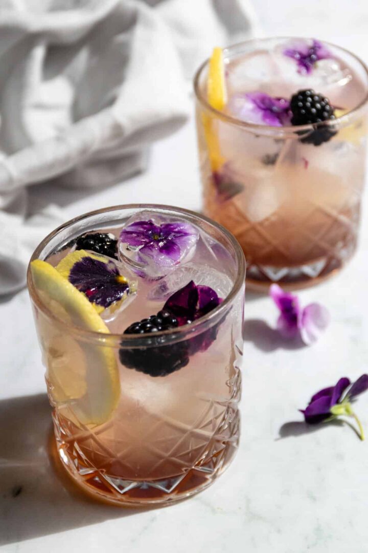 two glasses with bramble cocktail, wildflower ice cubes, lemon slice and blackberries
