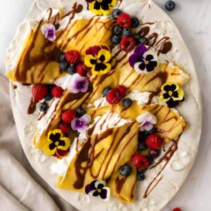 a large plate with crepes and whipped cream inside the crepes. The crepes are garnished with berries, chocolate sauce, and flowers. A kitchen towel on the side.