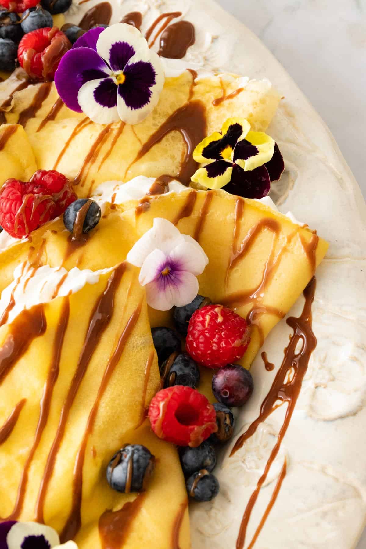 a plate with crepes and whipped cream inside the crepes. The crepes are garnished with berries, chocolate sauce, and flowers