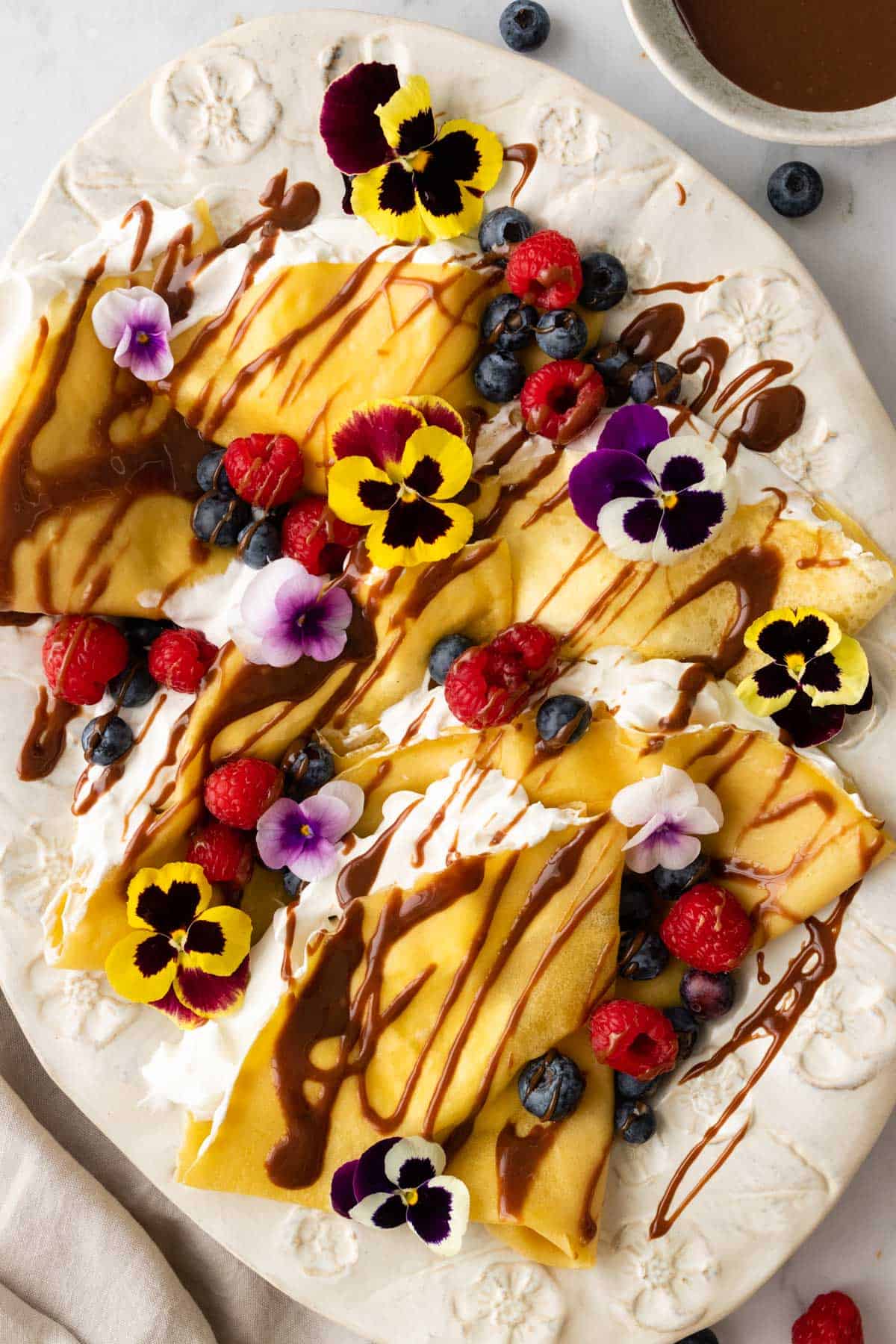 a large plate with crepes and whipped cream inside the crepes. The crepes are garnished with berries, chocolate sauce, and flowers