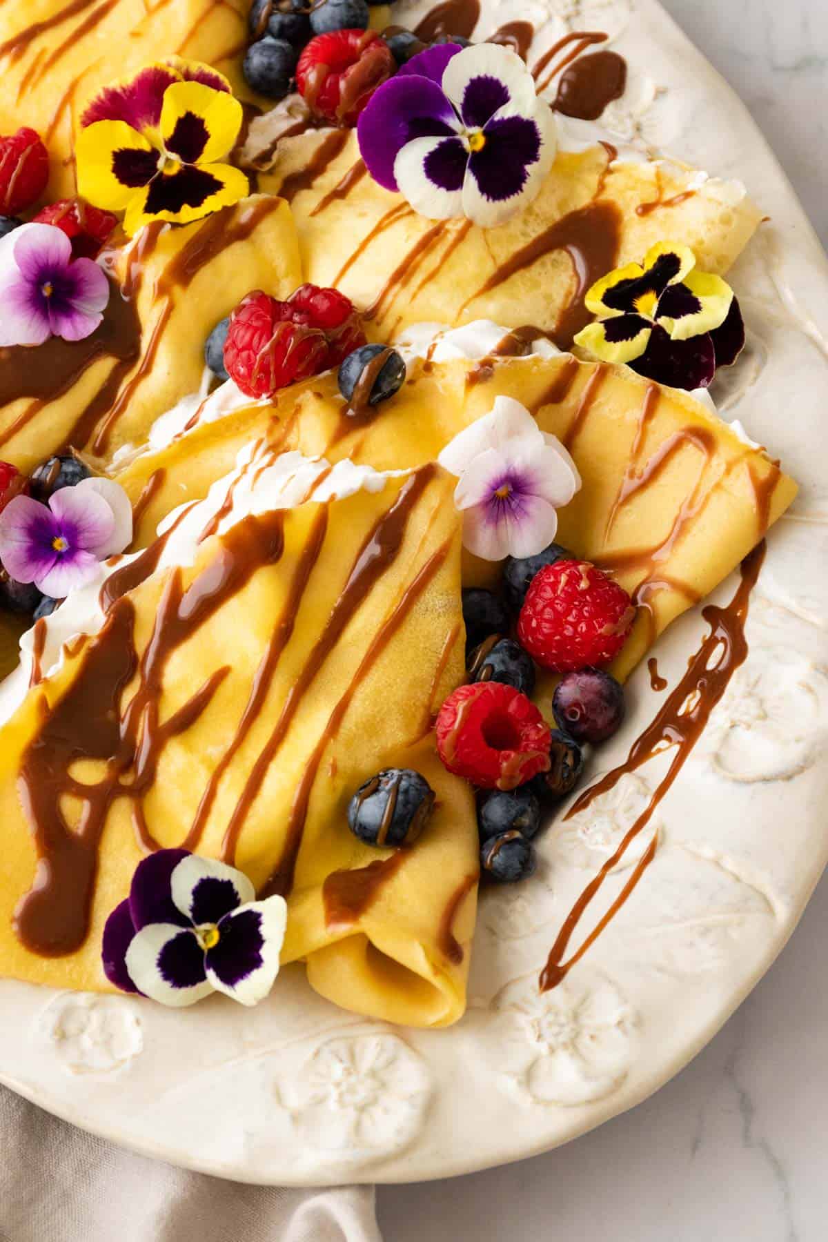 a large plate with crepes and whipped cream inside the crepes. The crepes are garnished with berries, chocolate sauce, and flowers