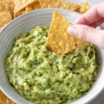one hand dipping a tortilla chip inside a bowl with guacamole