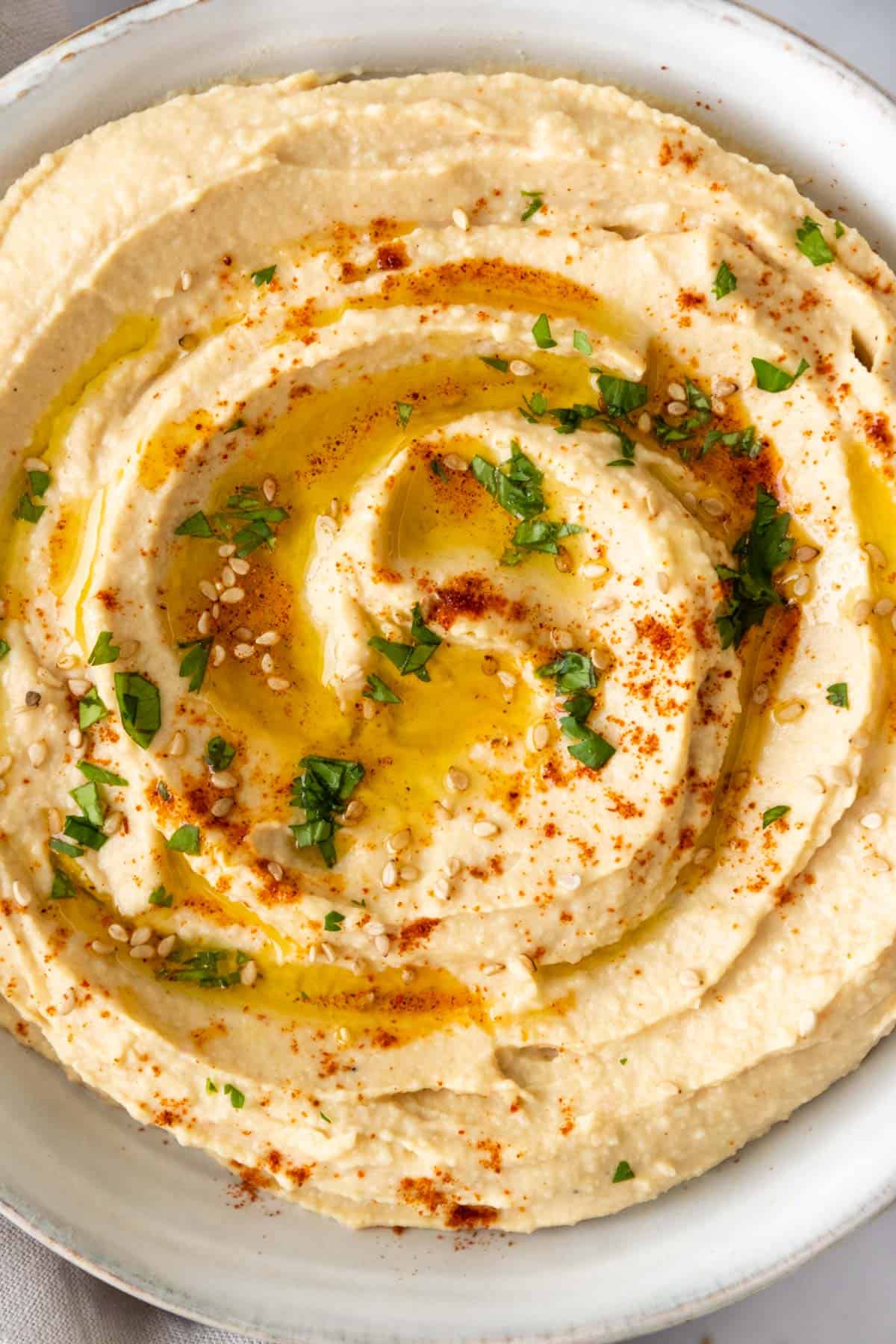 close up of a bowl with hummus garnished with sesame seeds, paprika, chopped parsley, and olive oil